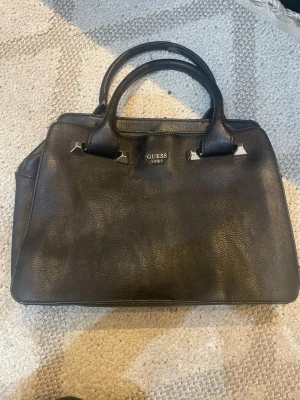 Svart handväska från Guess - Stilren svart handväska från Guess med två rejäla handtag och silverfärgade metalldetaljer. Väskan har en klassisk rektangulär form och är tillverkad i ett slätt konstläder. Perfekt för dig som gillar en enkel men snygg look. Sminkfläckar inuti men inget som stör. Äkta