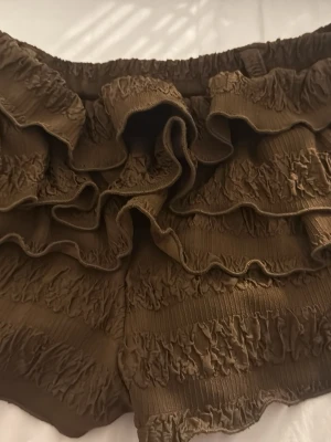 Bruna volangshorts med resårmidja - Supersöta bruna shorts med flera volanglager och smockdetaljer. Shortsen har elastisk midja för skön passform och är perfekta för varma dagar. Materialet känns mjukt och luftigt, vilket gör dem riktigt bekväma och lätta att bära.
