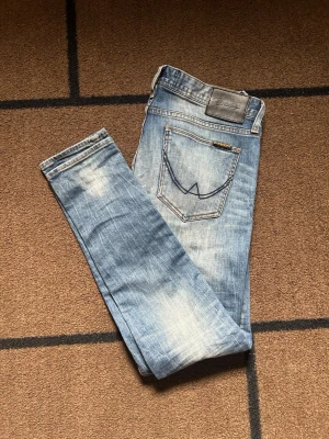 Ljusblå slim fit jeans Nudie Jeans - Snygga ljusblå slim fit jeans från Nudie Jeans med klassisk femficksdesign och tydliga slitningar på benen. Jeansen har en broderad bakficka och läderpatch i midjan. Perfekta för en avslappnad och trendig look.