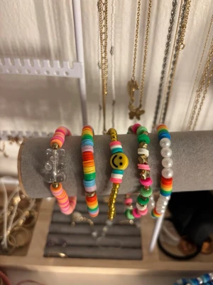 Färgglada pärlarmband set - Säljer ett set med fem färgglada armband i olika stilar. Armbanden har pärlor i regnbågens färger, guldiga detaljer, ett smileyface och vita plastpärlor. Perfekta för att ge din outfit en lekfull och trendig touch. Alla armband är elastiska och passar de flesta handleder.
