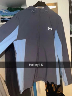 Blågrå hoodie från Under Armour - Snygg blågrå hoodie från Under Armour med ljusblå detaljer längs sidor och ärmar. Dragkedja framtill, justerbar huva med snören och logga på bröstet. Tillverkad i ett mjukt och funktionellt material som passar perfekt till träning eller chill.
