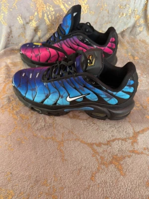 Nike Air Max Plus blå/rosa sneakers - Nike Air Max Plus sneakers med gradient färgskiftning från blått till rosa. Skorna har svarta detaljer, vit swoosh och robust svart sula med gula och röda inslag undertill. Ovandelen är i syntetmaterial med typiska TN-loggor och vågiga linjer. Perfekta för dig som gillar färgstarka sneakers.