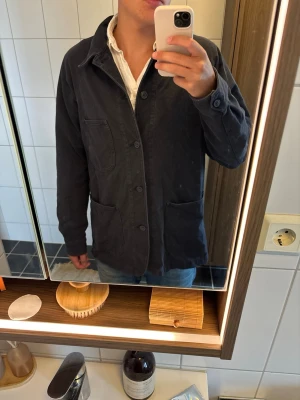 Mörkblå overshirt med stora fickor - Säljer en mörkblå overshirt i bomull med avslappnad passform. Jackan har stora utanpåliggande fickor framtill, knäppning med fyra knappar och klassisk krage. Perfekt att slänga över en t-shirt eller skjorta för en snygg lager-på-lager-look.
