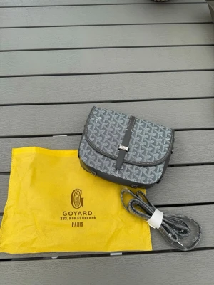 Grå axelväska från Goy4rd - Snygg grå axelväska från Goy4rd med klassiskt mönster och silverfärgad spänndetalj. Väskan har justerbar axelrem och lock med magnetlås. Tillverkad i canvas med läderdetaljer. Perfekt för dig som gillar exklusiva accessoarer.