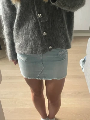 Ljusblå jeanskjol med slitningar - Trendig ljusblå jeanskjol i kort modell med råa kanter och slitna detaljer framtill. Kjolen har klassisk femficksdesign och dragkedja med knapp. Perfekt för dig som gillar en avslappnad och cool stil.