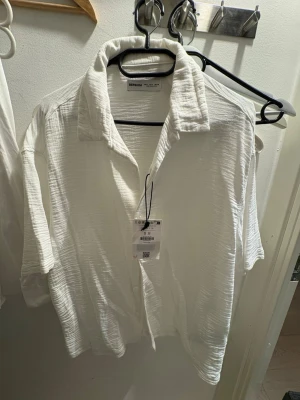 Vit skjorta från Bershka helt ny. - Säljer en vit skjorta från Bershka med avslappnad passform och korta ärmar. Skjortan har klassisk krage, knappar framtill och är tillverkad i ett lätt, strukturerat bomullstyg. Perfekt för en stilren och enkel look.