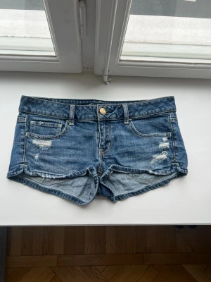 20s low waist jeansshorts - Säljer ett par blå denimshorts med slitna detaljer och råa kanter. Klassisk femficksmodell med knapp och dragkedja framtill. Shortsen har bälteshällor och kontrastsömmar. Perfekta för varma dagar! 20s! Storlek 6❤️❤️❤️