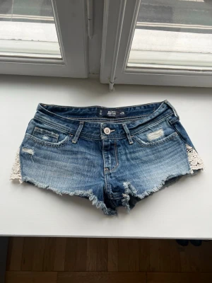 20s low waist jeansshorts med spetsdetaljer - Säljer ett par blå jeansshorts från Hollister med råa kanter och snygga slitningar. Shortsen har spetsdetaljer på sidorna och klassisk femficksdesign. Perfekta för sommaren och ger en avslappnad vibe. Från 20s