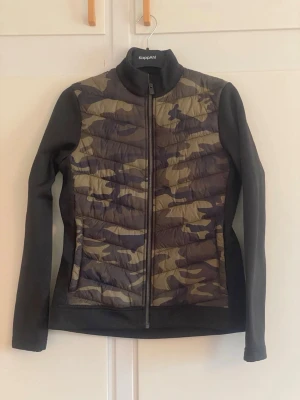 peakperformance camojacka med svarta ärmar - Snygg sportjacka från peakperformance med camouflage-mönstrad front och rygg i gröna och bruna toner. Svarta, stretchiga ärmar och sidor ger en skön passform. Jackan har hög krage och dragkedja framtill, perfekt för utomhusaktiviteter.
