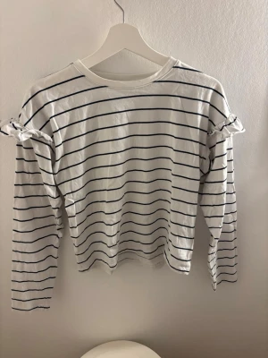Randig långärmad topp med volang - Vit långärmad topp med tunna svarta ränder och volangdetaljer på axlarna. Rund halsringning och rak passform. Perfekt för en avslappnad och trendig look.