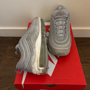 Nike Air Max 97 grå sneakers - Snygga Nike Air Max 97 sneakers i grått med ikoniska vågformade paneler och synlig Air-sula. Ovandelen är i mesh och syntetmaterial för en sportig look. Klassisk Nike-logga på sidan och tungan. Perfekta för dig som gillar streetwear och stilrena detaljer.