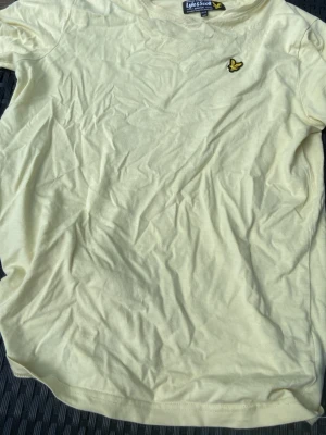 Ljusgul t-shirt från Lyle & Scott - Snygg ljusgul t-shirt från Lyle & Scott med klassisk rund hals och det ikoniska gula örnlogot på bröstet. Tillverkad i mjuk bomull som känns skön mot huden. Perfekt för en avslappnad och stilren look. Passar 13/12 åringar också beroende på kroppsbyggnad 
