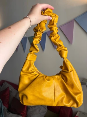 Gul scrunchie-handväska  - Trendig gul handväska med scrunchie-inspirerat handtag och justerbar axelrem. Väskan har en unik, rynkad design som sticker ut. Perfekt accessoar för att liva upp din outfit för sommaren. Inköpt på en marknad i Italien