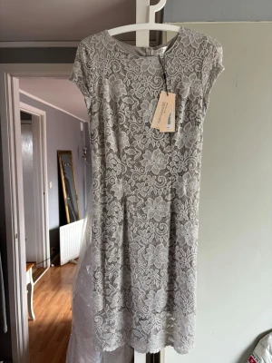 Ljusgrå spetsklänning med blommönster - Säljer en elegant ljusgrå spetsklänning med blommigt mönster och rund halsringning. Klänningen har korta ärmar och är tillverkad i ett tunt, spetsigt material som ger en romantisk känsla. Underklänning i stretchmaterial. Perfekt för sommarens alla tillfällen!