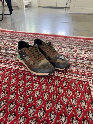Valentino rockrunners blå grön - Sneakers med camomönster i brunt, grönt och svart samt blå och turkosa detaljer på hälen. Skorna har låga skaft, svarta snören och en vit sula. Materialmix av mocka och textil ger en cool streetwear-look.