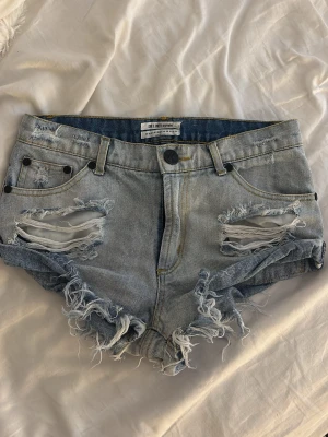 Ljusblå slitna jeansshorts One Teaspoon - Säljer ett par ljusblå jeansshorts från One Teaspoon med råa, fransiga kanter och stora slitningar framtill. Klassisk femficksmodell med knapp och dragkedja. Perfekta för sommaren och ger en avslappnad vibe. (XS-S)