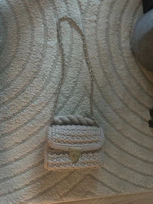 Beige virkad handväska med kedja - Snygg beige handväska i virkat material med guldfärgad kedjerem och tvinnat handtag. Väskan har ett lock med guldfärgat spänne och dekorativt emblem framtill. Perfekt accessoar för dig som gillar en unik och trendig look.