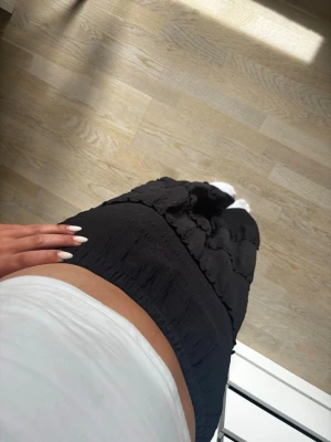 Svart långkjol med volangdetaljer - Säljer en svart långkjol med bred resår i midjan och flera volangdetaljer längs kjolen. Kjolen har en mjuk, något skrynklig struktur och faller snyggt längs benen. Perfekt för dig som gillar en enkel men ändå trendig look.