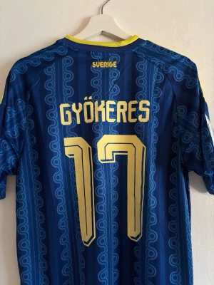 Blå Sverige fotbollströja Adidas - Säljer en blå Sverige fotbollströja från Adidas med gula detaljer och nummer 17 Gyökeres på ryggen. Tröjan har officiellt landslagsmärke, gula ränder på axlarna och retroinspirerat mönster. Perfekt för match eller träning.
