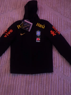 Nike Brasil GORE-TEX fotbollsjacka - Säljer en svart Nike GORE-TEX jacka med Brasilien fotbollslandslagets emblem på bröstet. Jackan har huva, dragkedja och tryck i gult och orange med sponsorloggor som Itaú och Vivo på ärmar och bröst. Perfekt för fotboll och träning utomhus.