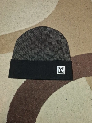 Mössa - Säljer en stilren svart och grå mössa med klassiskt rutigt mönster och vikt kant. Mössan har en snygg patch framtill och passar perfekt för dig som vill ha en clean och exklusiv look.