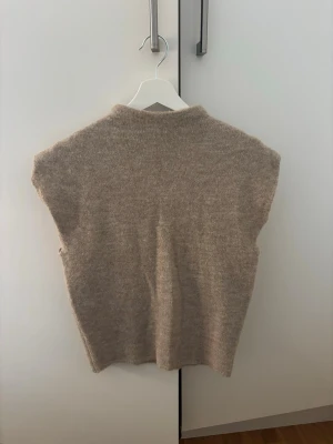 Beige stickad väst med hög hals - Mysig beige stickad väst med hög hals och breda ärmöppningar. Perfekt att slänga över en skjorta eller t-shirt för en avslappnad men ändå stilren look. Västen har en rak passform och är superenkel att matcha med olika outfits. Aldrig använd!