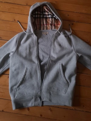 Grå zip hoodie från Burberry - Snygg grå hoodie från Burberry med dragkedja och klassiskt rutigt foder i huvan. Två stora fickor framtill och justerbar huva med snören. Perfekt för en chill och stilren look. Tillverkad i mjukt bomullsmaterial.