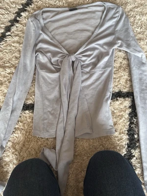 Ljusgrå långärmad topp med knyt - Supersnygg ljusgrå långärmad topp med djup v-ringning och knyt framtill. Sitter figurnära och har en croppad känsla. Perfekt att styla med jeans eller kjol för en trendig look.