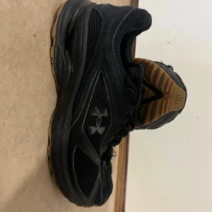 Svarta Under Armour sneakers - Snygga svarta sneakers från Under Armour med meshpaneler för extra andningsförmåga och detaljer i mocka. Skorna har en låg siluett, svart sula med gummidetaljer och diskret logga på sidan. Perfekta för en sportig och stilren look.