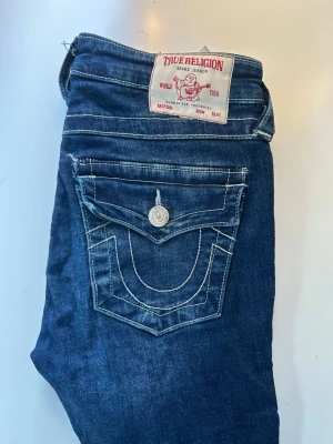 Blå bootcut jeans från True Religion - Säljer ett par blå bootcut jeans från True Religion med klassiska kontrastsömmar och fickor med lock och knappar bak. Jeansen har råa, fransiga benslut och en ikonisk patch bak i midjan. Perfekta för dig som gillar en avslappnad men trendig look.