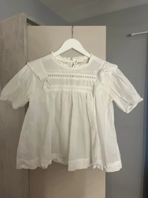 Vit blus med volang/bluzka - Söt vit blus från Gina Tricot med korta ärmar, volangdetaljer och broderade partier framtill. Blusen har en rund halsringning med liten volangkant och knäppning bak i nacken. Perfekt för en trendig och romantisk look.