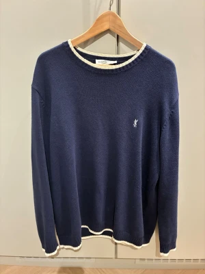 Mörkblå stickad tröja från Yves Saint Laurent Laurent - Mörkblå stickad tröja från Yves Saint Laurent med vit kant vid hals, ärmslut och nederkant. Tröjan har rund halsringning och en liten broderad logga på bröstet. Perfekt för dig som gillar klassisk och stilren design.