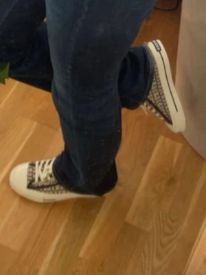 Beige och svarta Guess sneakers - Snygga sneakers från Guess i beige och svart med tydligt logomönster över hela skon. Klassisk låg modell med vita snören och vit sula. Perfekta för dig som vill ha en trendig och stilren look med tydlig branding.