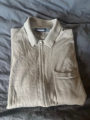 Beige ziptröja från J.Lindeberg 100% merino ull - Stilren beige ziptröja från J.Lindeberg 100% merino ull med krage och bröstficka. Tröjan har långa ärmar med ribbade muddar och är tillverkad i ett tunt, stickat material. Perfekt för lager-på-lager och enkel att matcha med andra plagg. Passar även S
