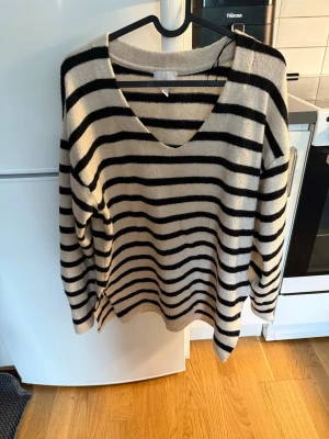 Randig stickad tröja med v-ringning - Beige stickad tröja med svarta horisontella ränder och djup v-ringning. Tröjan har lång ärm och avslappnad passform, perfekt för lager-på-lager. Mjuk och skön känsla, passar till jeans eller kjol.