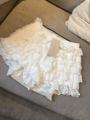 Nya i förpackning! Vita volangshorts med spets från Muséra - Superfina vita shorts från Muséra med flera lager volanger och spetsdetaljer. Perfekta för dig som vill ha en söt och trendig look. Shortsen har en feminin och lekfull stil med hög midja och mjukt material.