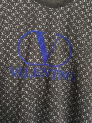 Svart Valentino tröja med logga - Cool svart tröja från Valentino med all-over V-mönster och stor blå logga på bröstet. Tröjan har rund halsringning, långa ärmar och ribbade muddar. Perfekt för dig som gillar streetwear och vill sticka ut med en designerpiece.