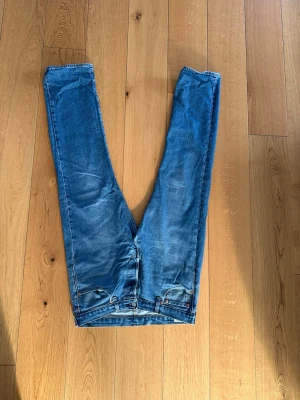Blå raka jeans från Levi's - Säljer ett par klassiska blå jeans från Levi's med raka ben och femficksmodell. Jeansen har en snygg tvättad look och detaljer som bruna sömmar och läderpatch bak i midjan. Perfekta för en avslappnad stil.