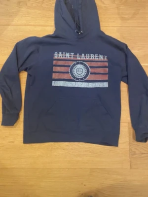 Blå hoodie från Saint Laurent - Snygg blå hoodie från Saint Laurent med tryck framtill i vitt och rött samt logga. Klassisk huva med snörning och känguruficka. Perfekt för en chill och trendig look. Modellen är lite liten i passform! Hör gärna av er kring frågor eller funderingarna