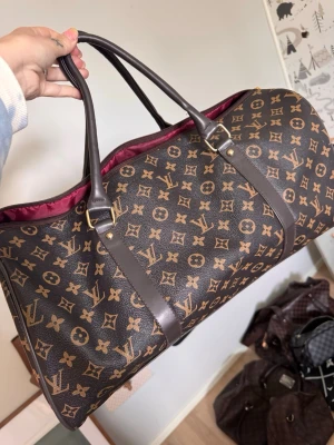  weekendbag - Snygg weekendbag  i klassiskt brunt monogrammönster med mörkbruna läderdetaljer och handtag. Väskan har guldfärgade metalldetaljer och vinrött foder. Perfekt för resor när du vill ha både stil och funktion.