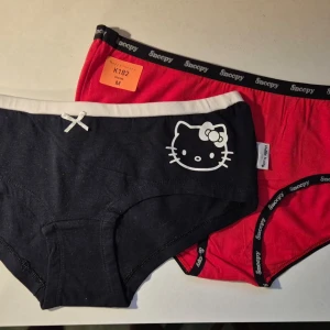 Hello Kitty och Snobben trosor storlek M - Set med två trosor i storlek M. Dels ett par söta svarta trosor från Hello Kitty med vit kant och liten rosett framtill. På ena höften finns en vit Hello Kitty-logga. Bekväm midjemodell i mjukt bomullsmaterial, perfekt för dig som gillar gulliga detaljer. Dels ett par snygga röda trosor med svart kant och Snobben-tryck på fram och baksida. Oanvända, nya, ingen lapp kvar. Från restlager. Äkta Y2K.