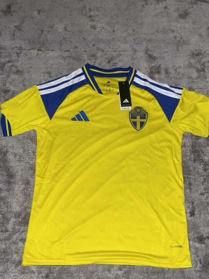 Sverige fotbollströja Adidas gul/blå - Gul fotbollströja från Adidas med blå och vita detaljer, Sveriges landslagsemblem på bröstet och 'Sverige' tryckt i nacken. Tröjan har korta ärmar, rund hals och är tillverkad i lätt funktionsmaterial. Perfekt för match eller träning.