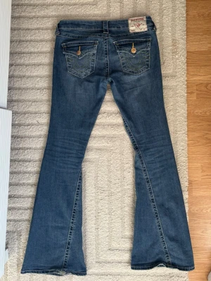 Blå bootcut jeans från True Religion - Säljer ett par blå bootcut jeans från True Religion med klassiska kontrastsömmar och snygga fickdetaljer bak med knappar. Jeansen har låg midja och är tillverkade i denim med lätt tvättad look. Perfekta för dig som gillar retrovibbar och markerad siluett.
