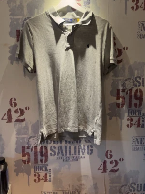 Grå pikétröja från Polo Ralph Lauren - Klassisk grå pikétröja från Polo Ralph Lauren i slim fit-modell. Tröjan har korta ärmar, krage med knappar och den ikoniska broderade loggan på bröstet. Perfekt för en stilren och avslappnad look.