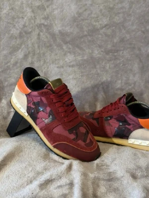 Valentino röda camo sneakers - Snygga sneakers från Valentino i röd och vinröd camo med svarta och lila detaljer. Skorna har orangeröd häl, svarta stjärnor och beige partier med nitar. Ovandelen är i mocka och textil, yttersulan är beige med svarta detaljer. Perfekta för dig som vill sticka ut. Obs endast skorna ingår