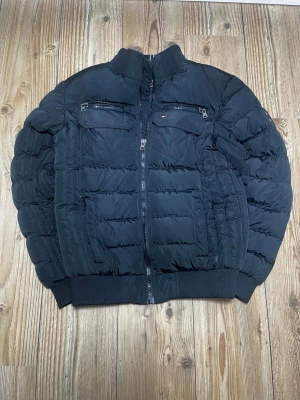 Svart pufferjacka Tommy Hilfiger - Svart pufferjacka från Tommy Hilfiger med hög krage, två bröstfickor med dragkedja och ribbade muddar. Jackan har klassisk quiltad design och diskret logga på bröstet. Perfekt för kalla vinterdagar och riktigt snygg streetstil.