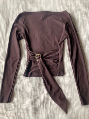 Brun långärmad topp med knyt - Snygg brun långärmad topp med bred båturringning och knytband i midjan med metallspänne. Toppen har en figurnära passform och är tillverkad i ett mjukt, stretchigt material som känns bekvämt mot huden.