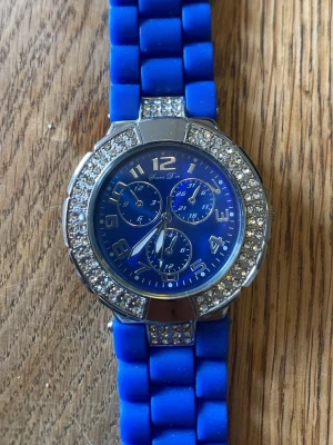Blå klocka med strassdetaljer - Snygg blå klocka från Geneva med glittrande strass runt urtavlan och matchande blått armband i syntetmaterial. Urtavlan är blå med silverfärgade siffror och flera dekorativa små urtavlor. Perfekt accessoar för att lyfta din stil.