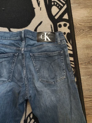 Blå raka jeans från Calvin Klein - Snygga blå jeans från Calvin Klein med rak passform och klassisk femficksdesign. Jeansen har en lätt tvättad look och silverfärgade nitar. Perfekta för en avslappnad stil och passar till det mesta.