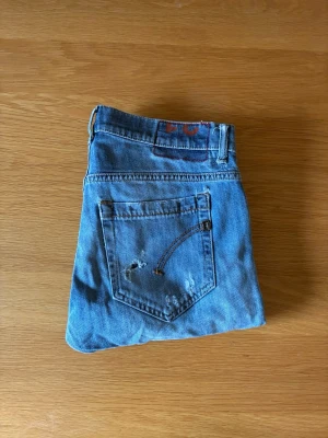 Dondup George jeans  - Säljer ett par klassiska blå dondup jeans med raka ben och snygga slitningar på bakfickan. Jeansen har en enkel design med fem fickor och synliga sömmar. Perfekta för en avslappnad och trendig look.